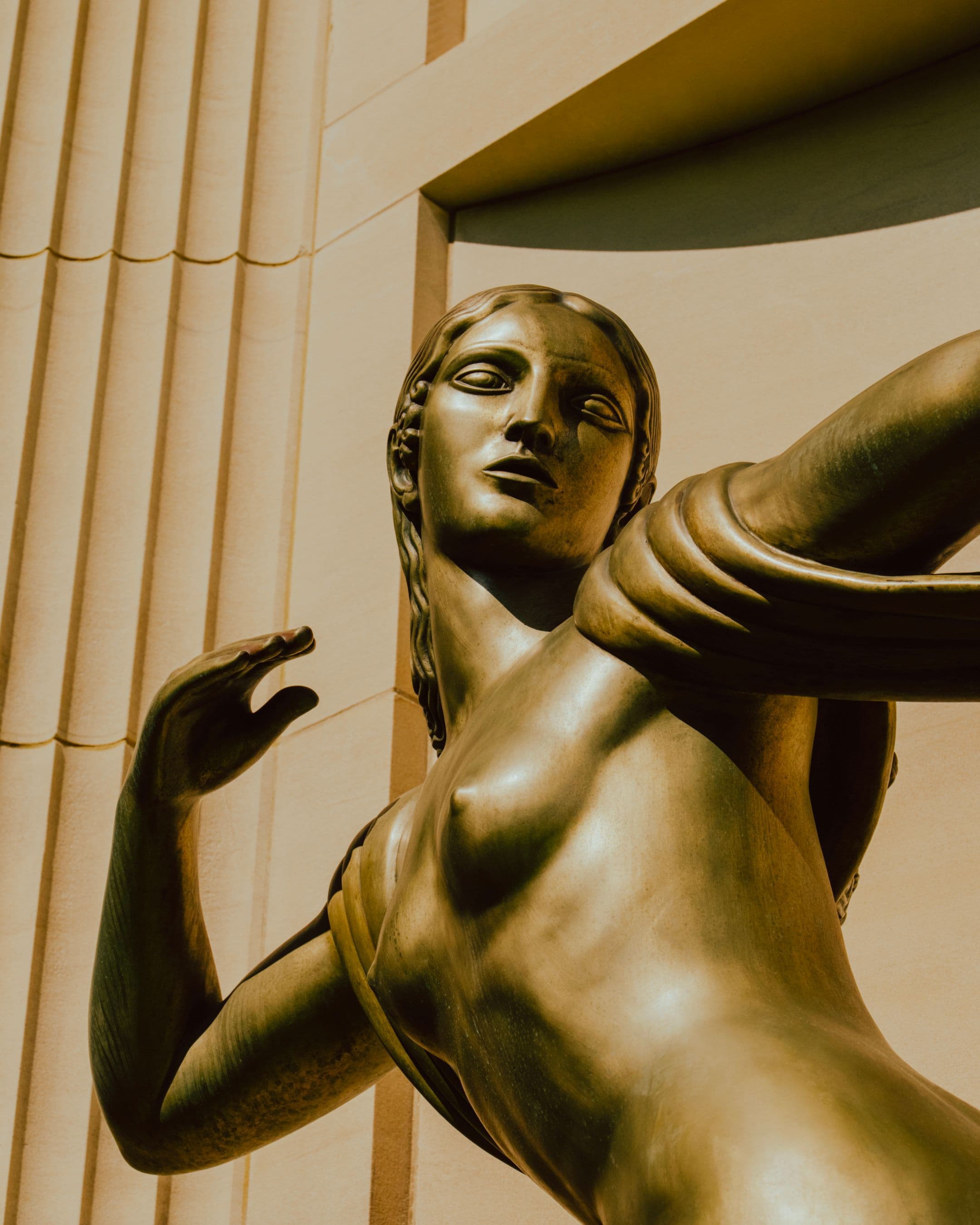 A close-up, low-angle view of a bronze figurative sculpture of a woman with arms outstretched, photographed against the stone columns of the Norton Museum of Art's exterior.