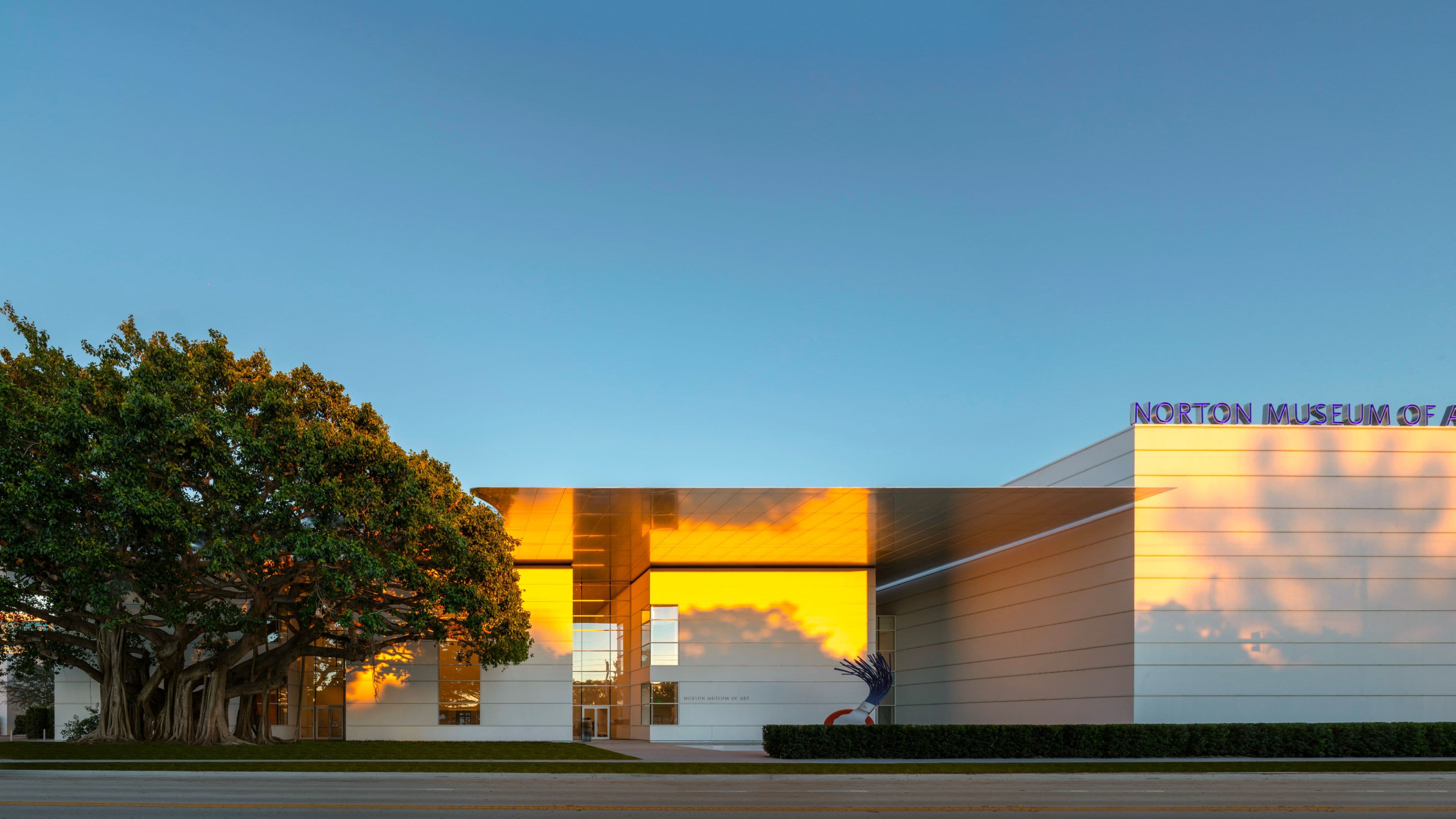 The exterior of the Norton Museum of Art at golden hour, with warm sunset light reflecting off the building's modern facade. A large banyan tree frames the left side of the building, and a blue sculptural artwork is visible near the entrance.