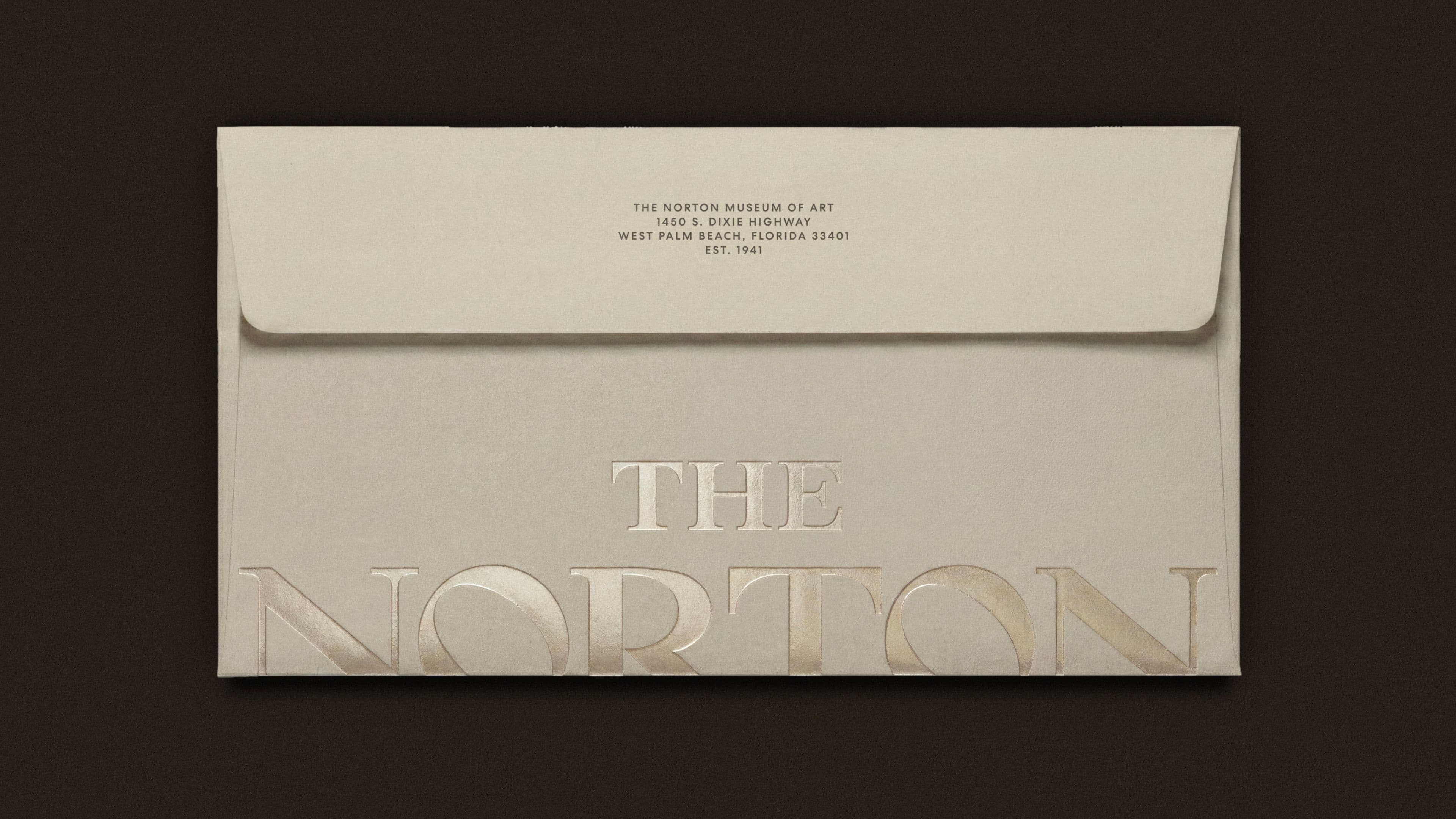 A cream-colored envelope with the Norton Museum of Art's return address printed at the top, and "The Norton" embossed in large elegant lettering across the front, set against a dark brown background.