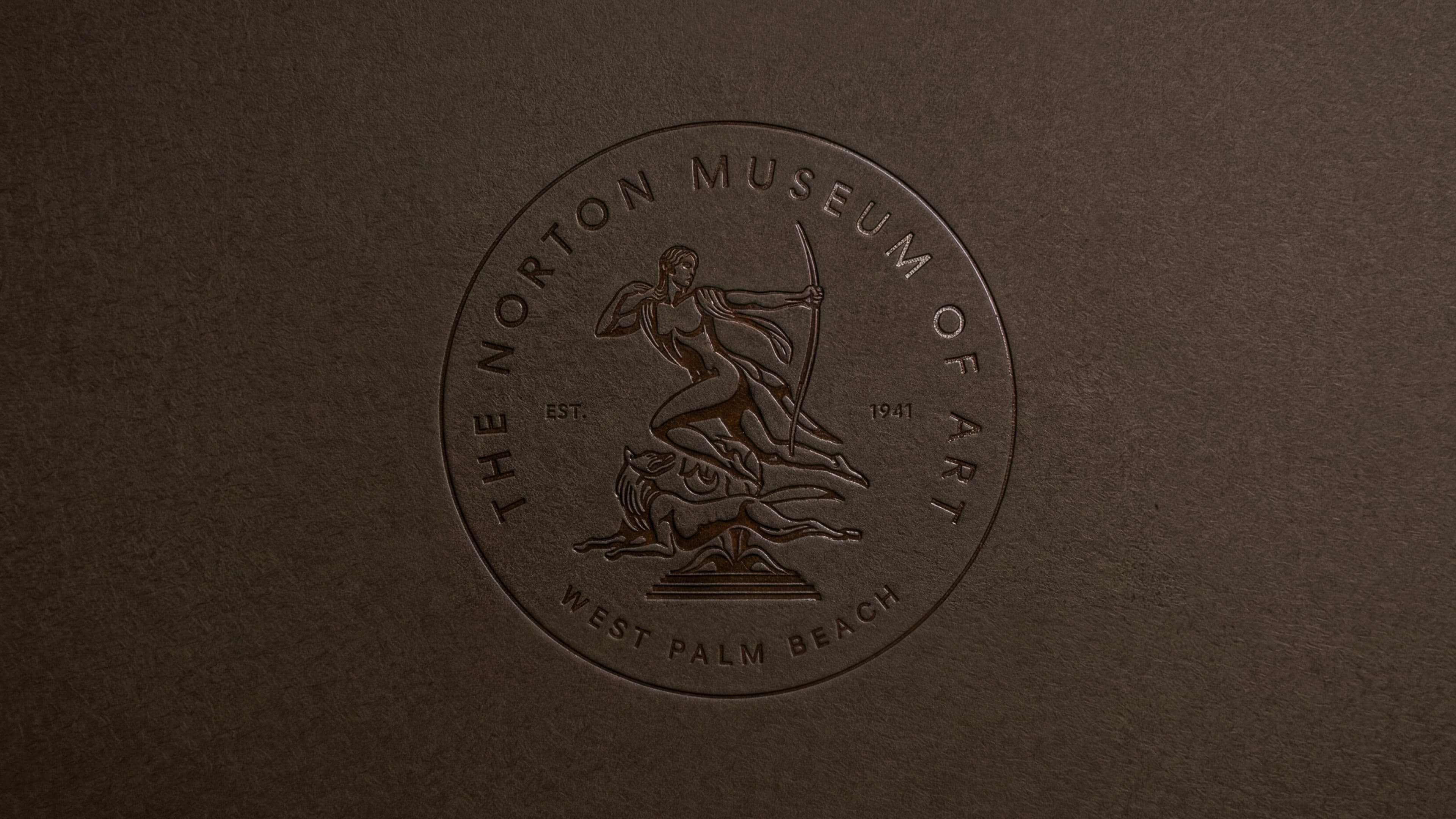 The Norton Museum of Art's circular seal embossed on dark brown textured paper, featuring a classical figural illustration and the text "The Norton Museum of Art, Est. 1941, West Palm Beach."